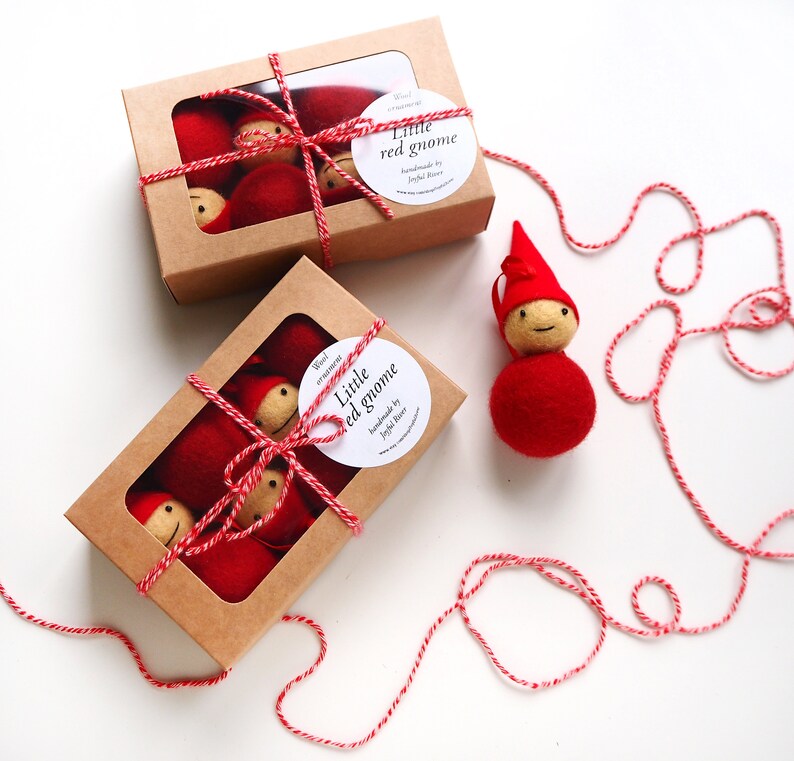 May include: Two cardboard boxes with clear windows, each containing several red gnome ornaments. The boxes are tied with red and white twine. A single gnome ornament sits beside the boxes. The ornaments have red bodies and hats, with beige faces. The text "Little red gnome" is on a white sticker.