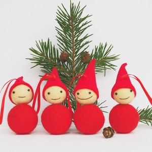 May include: Four Christmas ornaments shaped like smiling gnomes, each with a red body and hat, and a light-colored head. They have red ribbon loops for hanging. A sprig of evergreen and a pine cone are in the background.