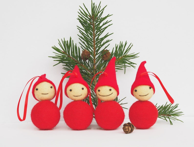 May include: Four Christmas ornaments shaped like smiling gnomes, each with a red body and hat, and a light-colored head. They have red ribbon loops for hanging. A sprig of evergreen and a pine cone are in the background.