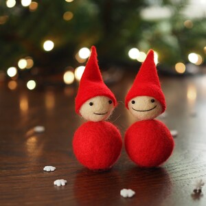 May include: Two handmade Christmas gnome ornaments. Each gnome has a red conical hat, a round red body, and a cream-colored head with a smiling face. The ornaments are on a wooden surface with snowflake decorations and a blurred background of Christmas lights.