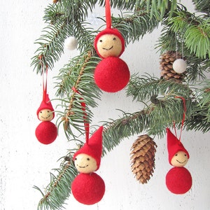 May include: Christmas tree ornaments featuring red felt gnome-like figures with smiling faces and pointed hats. The ornaments are hanging from a pine branch, alongside a pine cone and white pom-poms. The ornaments are suspended by red ribbons.
