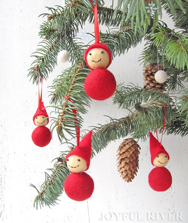 May include: Christmas tree ornaments featuring red felt gnome-like figures with smiling faces and pointed hats. The ornaments are hanging from a pine branch, alongside a pine cone and white pom-poms. The ornaments are suspended by red ribbons.
