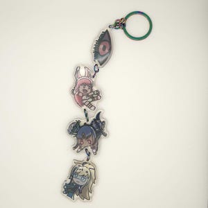 May include: A keychain featuring four anime-style charms. The charms include a large eye, a pink-haired character, a dark-haired character, and a blonde character. The keychain has a rainbow-colored ring and connecting links, all on a white background.