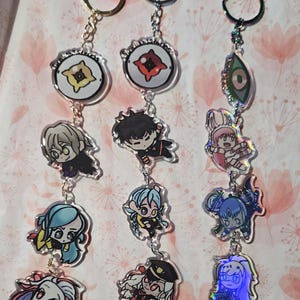 Puede incluir: Tres llaveros con diseños de personajes de dibujos animados. Cada llavero tiene una parte superior circular con un anillo dorado o plateado, una cadena y múltiples colgantes acrílicos. Los colgantes presentan ilustraciones coloridas de estilo anime.