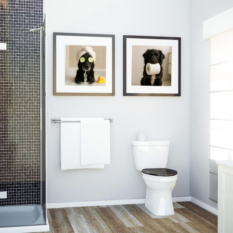 May include: Two framed prints of black dogs in a bathroom setting. One dog wears a spa towel and cucumber slices, the other holds toilet paper. A white toilet, towels, and shower are also visible.