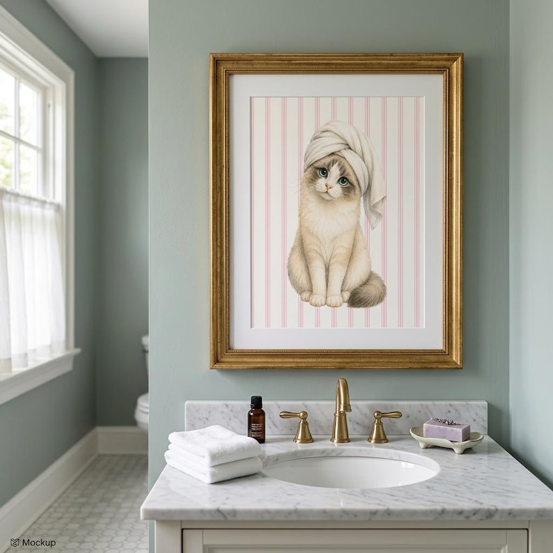 K&ouml;nnte beinhalten: Ein gerahmter Druck mit einer Katze, die ein Handtuch auf dem Kopf tr&auml;gt, vor einem rosa-wei&szlig; gestreiften Hintergrund. Das Kunstwerk befindet sich in einem goldenen Rahmen und h&auml;ngt &uuml;ber einem Badezimmerwaschbecken. Die Katze hat blaue Augen und einen flauschigen Schwanz.