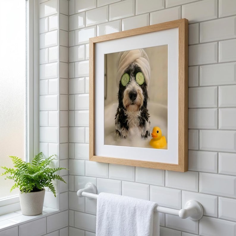May include: A framed print shows a black and white dog in a bathtub with cucumber slices on its eyes and a towel on its head. A yellow rubber duck sits in the tub. The print is in a light brown wooden frame, hanging on a white tiled wall.