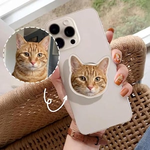 May include: A white phone case featuring a tabby cat face design. The case has a pop-up grip with the same cat face. The image also shows a close-up of the cat's face in a circle.