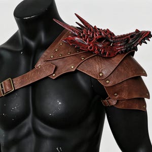 May include: A brown leather-like shoulder pauldron featuring a layered plate design secured with metallic studs and buckles. A sculpted, three-dimensional red and black dragon head ornament is attached to the top of the shoulder piece.