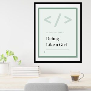 May include: A framed print with the text "Debug Like a Girl" in a modern font. The print has a light green background with coding symbols and a black frame. A stack of white books, a small plant, and a cup of coffee are on the desk.