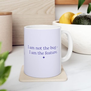 May include: White ceramic mug with the text "I am not the bug - I am the feature" in purple. The mug sits on a beige coaster. The mug has a white handle and is on a white surface.