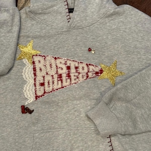 May include: A gray hooded sweatshirt with a Boston College pennant design. The pennant is red and white plaid with the words "BOSTON COLLEGE". Gold star and small decorative pins are also on the sweatshirt.