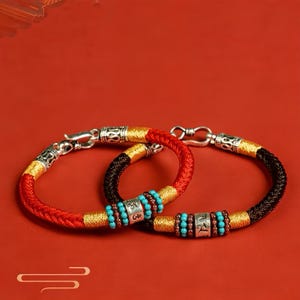 May include: Two braided bracelets. One is red and gold, the other is black and gold. Both have silver clasps and decorative silver beads with turquoise and brown beads. The bracelets are displayed on a red background.