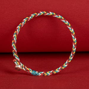 May include: A braided bracelet with a colorful pattern of red, white, yellow, blue, and black threads. The bracelet has a small silver bell charm attached. The background is a solid red color.