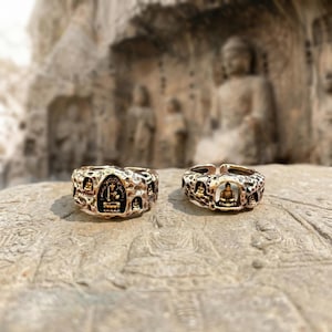 Puede incluir: Dos anillos de color bronce con una superficie texturizada. Cada anillo presenta una figura central de Buda y figuras de Buda más pequeñas alrededor de la banda. Los anillos están sobre una superficie de piedra con un fondo borroso.