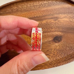 Puede incluir: Un anillo plateado con caracteres chinos rojos y dorados. El anillo se sostiene en una mano, sobre un fondo de madera. El diseño del anillo es detallado, con caracteres pintados a mano. Una joya.
