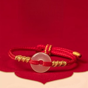 Könnte beinhalten: Rotes Armband mit einem kreisförmigen, durchscheinenden Stein. Das Armband hat goldfarbene Akzente und eine rote Kordel, vor rotem Hintergrund. Das Design ist schlicht und elegant.