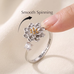 Könnte beinhalten: Ein silberfarbener Drehring mit einer texturierten Lotusblüte, goldenen Details in der Mitte und einem einzelnen klaren runden Stein. Der Ring ist mit dem Text You are my sun shine graviert, und das Bild enthält den Text Smooth Spinning.