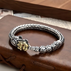 Könnte beinhalten: Silberfarbenes Armband mit geflochtenem Band und detailliertem goldfarbenem Löwenkopfverschluss. Das Armband hat eine Reihe von silbernen Perlen mit komplizierten Mustern. Ein kleiner roter Edelstein ist in das Auge des Löwen eingesetzt. Das Armband wird auf einer braunen Lederoberfläche präsentiert.