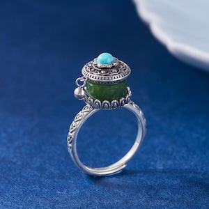 Könnte beinhalten: Silberring mit einem grünen Jadezylinder und einem türkisfarbenen Stein. Der Ring hat ein verziertes Design und einen kleinen Glockenanhänger. Der Ring ist vor einem blauen Hintergrund platziert.
