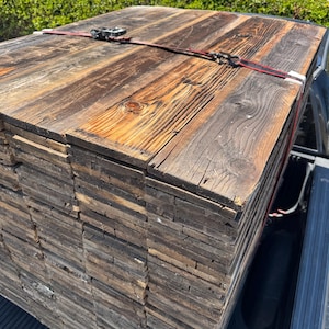 May include: A large stack of weathered, dark brown reclaimed wooden planks secured with a red and black ratchet strap. The boards feature visible wood grain, knots, and a rough, aged texture, stacked neatly in the bed of a pickup truck.