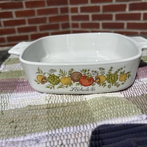 May include: White rectangular casserole dish with handles, decorated with a colorful vegetable and herb design. The dish has the word "L'Echalote" printed on the front. The dish is sitting on a multi-colored woven placemat.