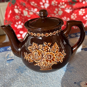May include: A brown ceramic teapot with a lid and handle. The teapot features a floral design in a lighter brown color. The teapot is sitting on a blue and white patterned tablecloth.
