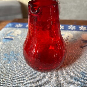 May include: A vibrant red glass pitcher with a crackle finish. The pitcher has a bulbous base and a slender neck with a pouring spout. The glass appears to be translucent, allowing light to pass through. The pitcher is likely used for serving beverages.