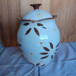 May include: A white ceramic jar with a lid, adorned with brown floral patterns and small yellow and red flower accents. The jar has a brown rim and a brown knob on the lid. The jar is sitting on a blue and white striped cloth.