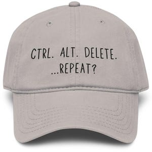 May include: A light gray baseball cap featuring the black text "CTRL. ALT. DELETE. ...REPEAT?" embroidered on the front. The cap has a structured crown, a curved brim with visible stitching, and a small button at the top center of the fabric dome.