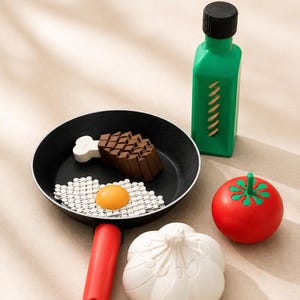 May include: A black frying pan with a red handle containing a brown toy meat piece with a white bone and a white toy fried egg with a yellow yolk. Beside the pan is a green bottle, a red tomato with a green stem, and a white garlic bulb.