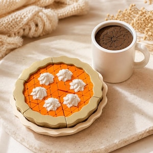 Puede incluir: Un pastel de juguete de madera con relleno naranja brillante, corteza festoneada color canela y seis copos blancos de crema batida, acompañado de una taza de cerámica blanca con un inserto circular marrón oscuro con un patrón geométrico.