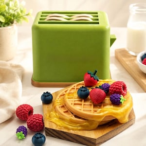 May include: A bright green toy toaster sits behind a wooden board holding two round waffles topped with red strawberries, purple blackberries, and raspberries, all covered in a thick, glossy, golden syrup, with loose berries scattered nearby.