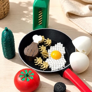 May include: A toy kitchen set featuring a black frying pan with a red handle containing a plastic fried egg, a brown chicken leg, and tan pasta shapes. Accessories include a green bottle, a green cucumber, a red tomato, and two white egg shells.