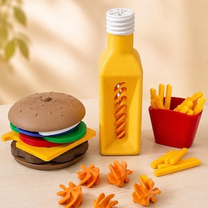 Op de afbeelding: Een speelgoedset bestaande uit een bruine hamburger met gestapelde groene, rode en gele lagen, een gele fles met een witte dop, een rode houder gevuld met gele frietjes en vijf kleine oranje stervormige onderdelen op een ondergrond.