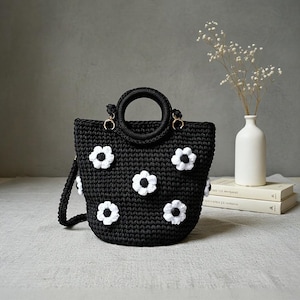 May include: A black crochet handbag with white daisy appliqués. The bag features a circular handle and a detachable shoulder strap. The bag is decorated with white daisy flowers and gold-colored hardware.