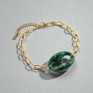 Può includere: Bracciale a catena dorata con un elemento centrale ovale verde marmorizzato. Il bracciale ha una chiusura a moschettone e una catena regolabile. L'elemento centrale ovale ha due ritagli circolari e una finitura lucida. Il bracciale è su una superficie grigia.