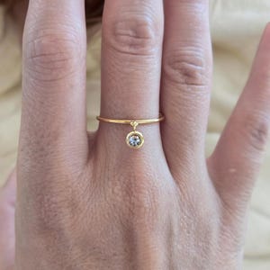 Peut inclure: Une fine bague dorée ornée d'un délicat pendentif circulaire suspendu. Le pendentif est serti d'une petite pierre transparente ronde à facettes qui capte la lumière, créant un éclat subtil sur l'anneau en métal poli.