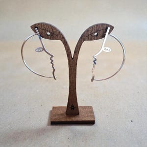 Peut inclure: Boucles d'oreilles créoles argentées avec un design de visage minimaliste, présentées sur un présentoir en bois. Les boucles d'oreilles présentent un profil avec un œil et une bouche, créant un look artistique unique. Le présentoir est en bois foncé.