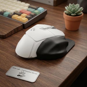 May include: A white computer mouse with a black ergonomic side grip attachment. The mouse features a prominent G logo on the top shell. A small silver rectangular plaque nearby displays the text Grip Lab - Contoured Series with a hand icon.