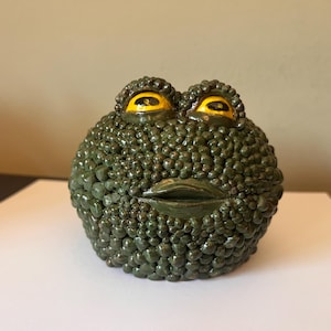 May include: A round, dark green ceramic frog figurine with a highly textured, bumpy surface. The frog features two prominent, glossy yellow eyes with black pupils and a wide, horizontal mouth opening centered on the front of the textured body.