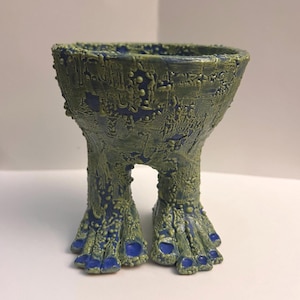 May include: A ceramic vessel with a wide, open top and a textured, moss-green exterior finish over a deep blue base. The container is supported by two thick, leg-like structures that terminate in wide, flat feet with distinct blue-painted toes.
