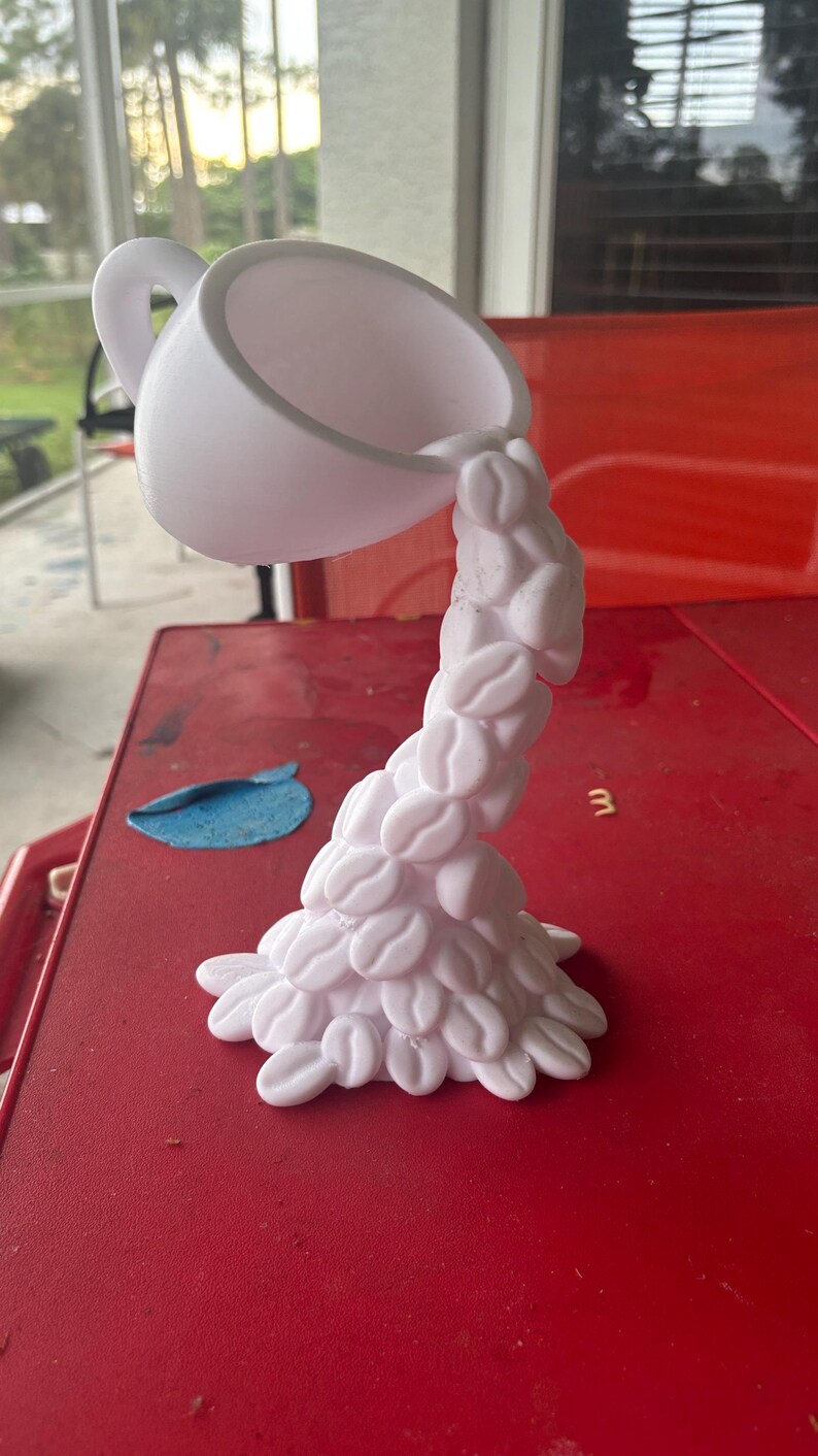 Peut inclure: Une sculpture de tasse &agrave; caf&eacute; blanche avec une anse, semblant verser des grains de caf&eacute;. Les grains forment une colonne en cascade, cr&eacute;ant une pi&egrave;ce d&eacute;corative unique. La sculpture est sur une surface rouge.