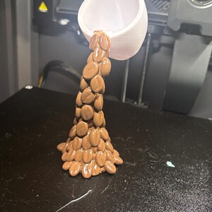Peut inclure: Une tasse blanche imprim&eacute;e en 3D semble verser un flux d'objets bruns de forme ovale. Les objets sont dispos&eacute;s pour ressembler &agrave; un &eacute;coulement de liquide. La tasse est positionn&eacute;e au-dessus d'une surface noire, probablement un plateau d'imprimante 3D.