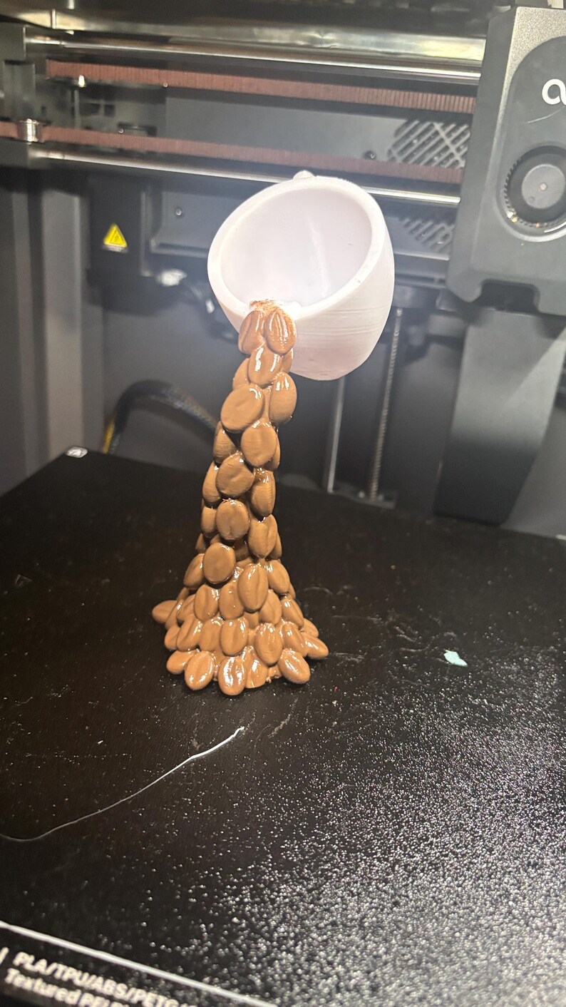 Peut inclure: Une tasse blanche imprim&eacute;e en 3D semble verser un flux d'objets bruns de forme ovale. Les objets sont dispos&eacute;s pour ressembler &agrave; un &eacute;coulement de liquide. La tasse est positionn&eacute;e au-dessus d'une surface noire, probablement un plateau d'imprimante 3D.
