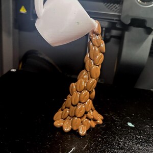 Peut inclure: Une sculpture imprim&eacute;e en 3D d'une tasse &agrave; caf&eacute; blanche versant un flux de grains de caf&eacute; bruns. Les grains forment une colonne en cascade, cr&eacute;ant une repr&eacute;sentation artistique du caf&eacute;. La sculpture est sur une surface noire.