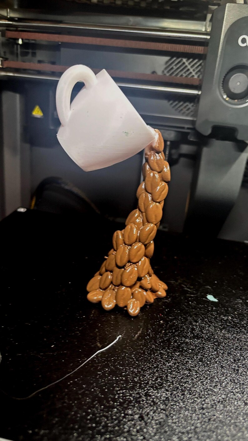 Peut inclure: Une sculpture imprim&eacute;e en 3D d'une tasse &agrave; caf&eacute; blanche versant un flux de grains de caf&eacute; bruns. Les grains forment une colonne en cascade, cr&eacute;ant une repr&eacute;sentation artistique du caf&eacute;. La sculpture est sur une surface noire.