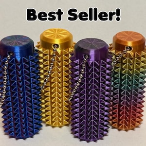 May include: Four colorful, cylindrical objects with a spiked texture and a small chain. The objects are blue, gold, purple, and rainbow-colored. The text "Best Seller!" is displayed above the objects.