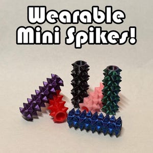 May include: A collection of cylindrical, hollow, spike-covered accessories in purple, red, black, pink, green, and blue. The text "Wearable Mini Spikes!" is printed in white with a black outline above the objects, which feature a textured, geometric surface.