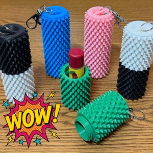 May include: A collection of colorful, textured lip balm holders with keychains. The holders are in various colors, including black, white, blue, pink, and green. One green holder contains a lip balm. A comic book style "WOW!" graphic is in the image.
